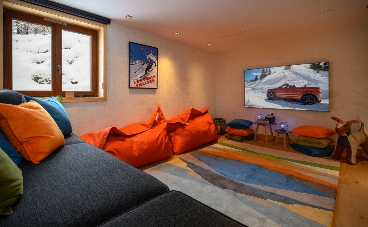 A cozy ski-themed living room with a dark gray sectional sofa, bright orange bean bags, a colorful striped rug, and a wall-mounted TV showing a snowy mountain scene.