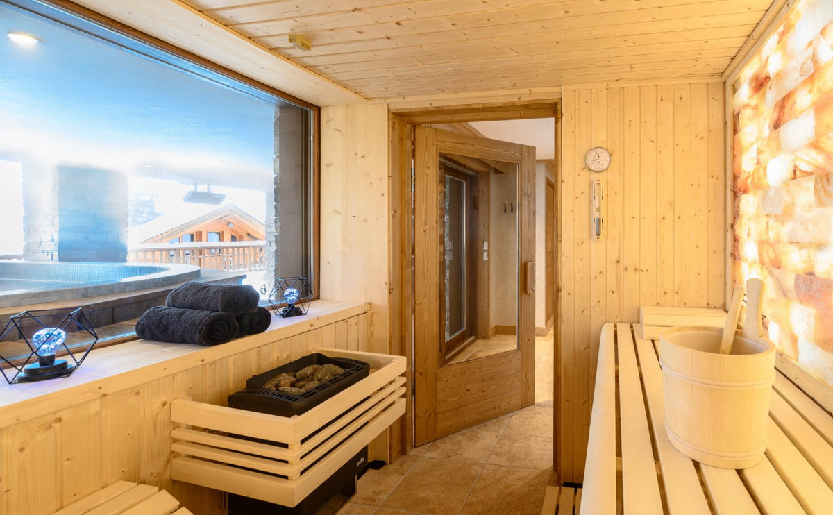 A warm, wood-paneled sauna room with a wooden bench, salt wall, steam table, towels, and a large window overlooking a snowy outdoor scene.