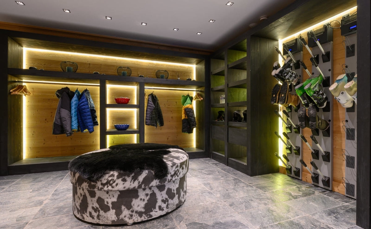 A modern ski shop interior with illuminated wood shelving, hanging jackets, wall-mounted skis, and a round black-and-white cowhide ottoman in the foreground.