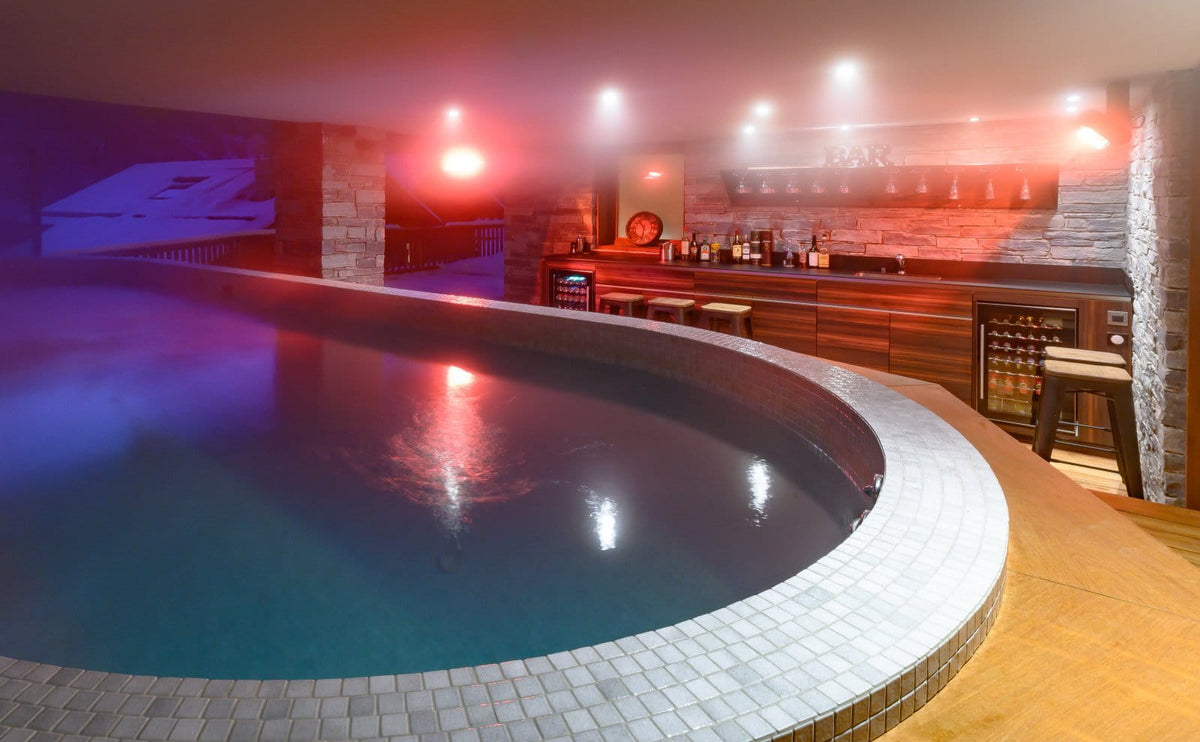 An indoor curved pool with blue water and warm red ambient lighting, featuring a tiled edge and a bar area in the background with stone walls and stools.