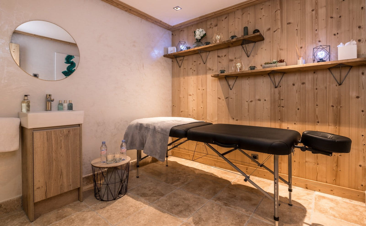 A spa treatment room featuring a black massage table, a wooden vanity cabinet with a white sink and round mirror, and a wood-paneled wall with floating shelves holding decorative items.