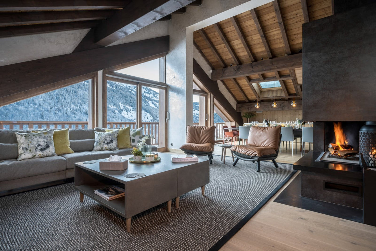A luxurious ski chalet living room featuring a large gray sectional sofa with green and yellow accent pillows, a low gray coffee table, two brown leather armchairs, a large black fireplace with a visible fire, and a high wood-beamed ceiling with panoramic windows overlooking a snowy mountain landscape.