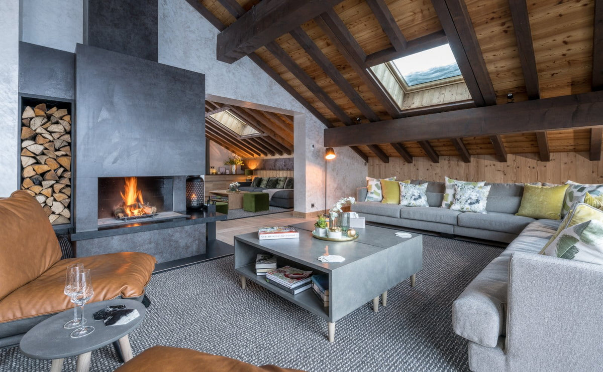 A modern mountain-style chalet living room featuring a large gray concrete fireplace with a visible fire, a gray sectional sofa with yellow and green pillows, a gray coffee table, and a brown leather armchair under a wooden-beamed attic ceiling.
