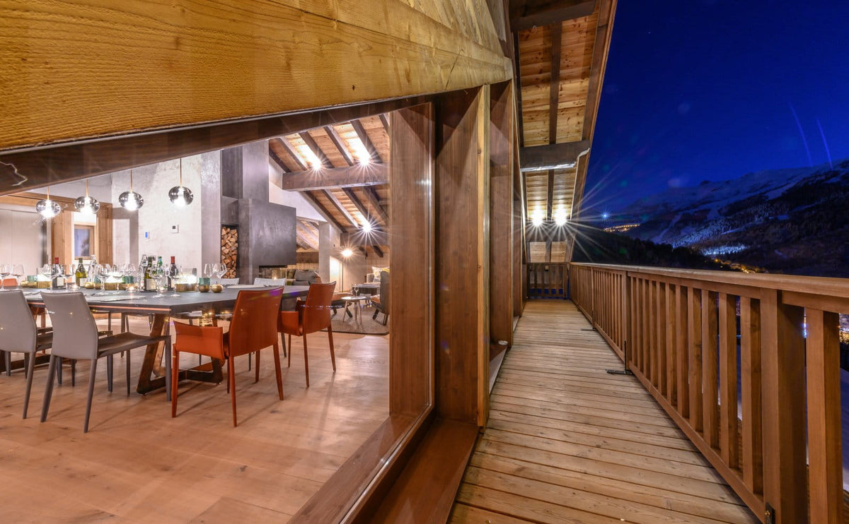 A modern wooden chalet balcony at night with a view of mountains in the distance, overlooking a dining area inside with wooden floors and chairs.