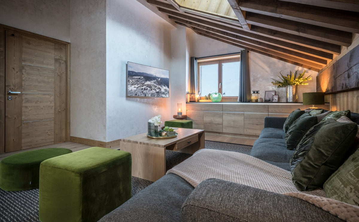 A modern, rustic-style living room featuring a gray sectional sofa with dark green pillows, two green velvet ottomans, a light wood coffee table, and a wall-mounted TV. The room has a sloped wooden ceiling with exposed beams, a window with warm lighting, and a textured plaster wall.