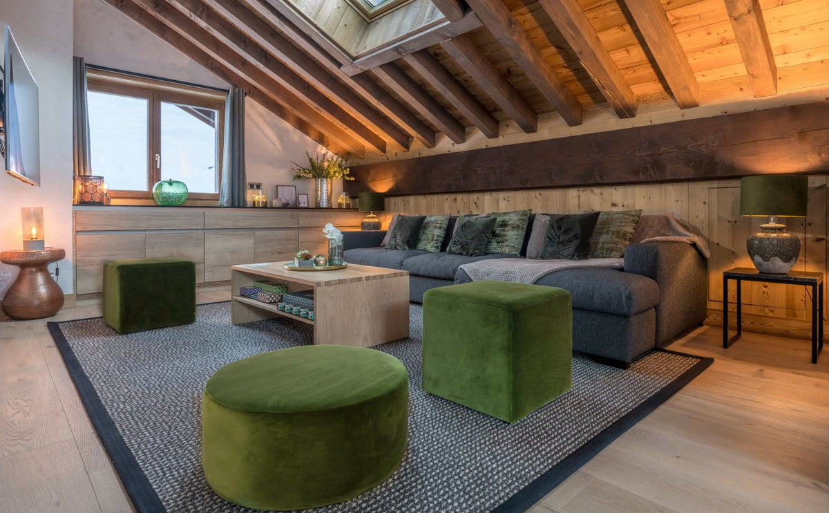 A cozy modern living room with a gray sectional sofa, two green velvet ottomans, a light wood coffee table, and a patterned rug, set against a rustic wood-beamed ceiling.