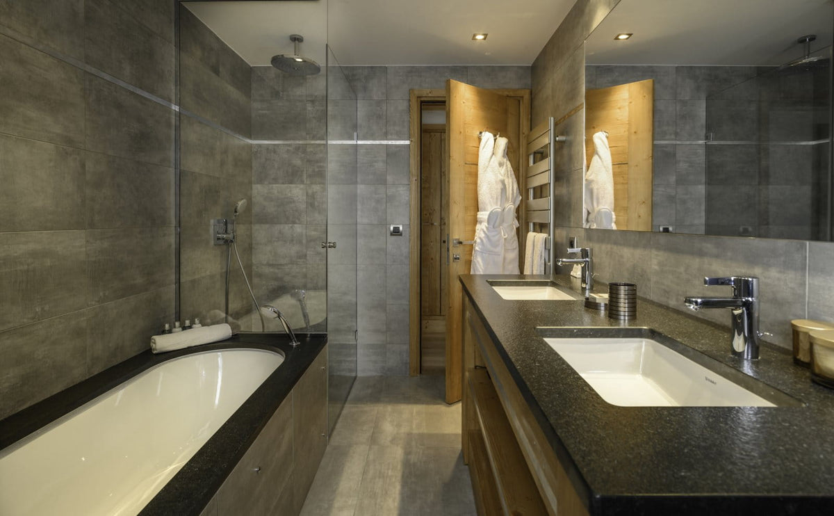 A modern bathroom interior with a long double vanity, white sinks, chrome faucets, dark stone countertops, gray tiled walls, a glass shower enclosure, and a bathtub along the left side.
