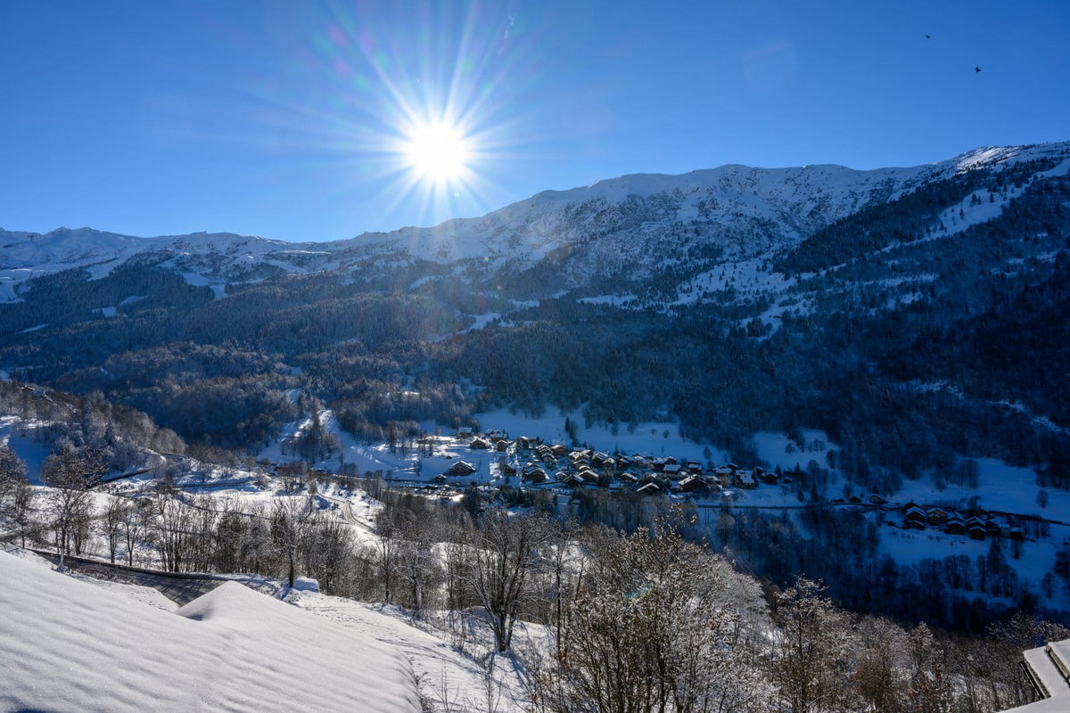 A bright winter mountain landscape with snow-covered peaks, a sunburst in the blue sky, a valley village, and a lake reflecting the snowy surroundings.