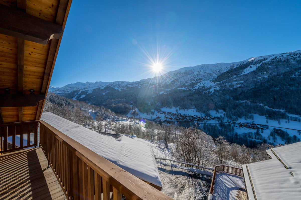 A sunny winter mountain landscape viewed from a wooden balcony, with snow-covered peaks, a clear blue sky, and bright sun rays shining over a valley.