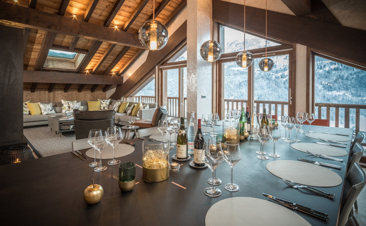 A luxury dining table set in a modern ski chalet with large windows overlooking a snowy mountain landscape. The table is styled with white plates, wine glasses, bottles, and gold candle holders, under spherical glass pendant lights.