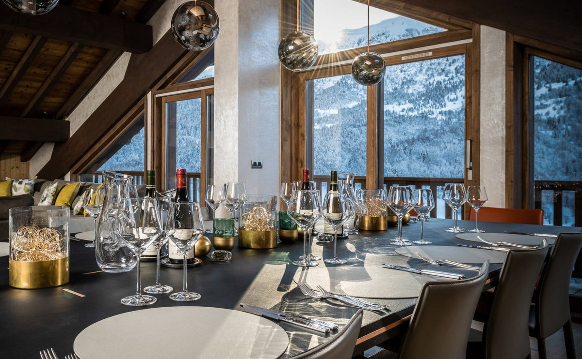 A luxury dining table set in a modern chalet with large windows overlooking a snowy mountain landscape. The table is elegantly arranged with wine glasses, plates, and gold and glass candle holders under spherical hanging lights.