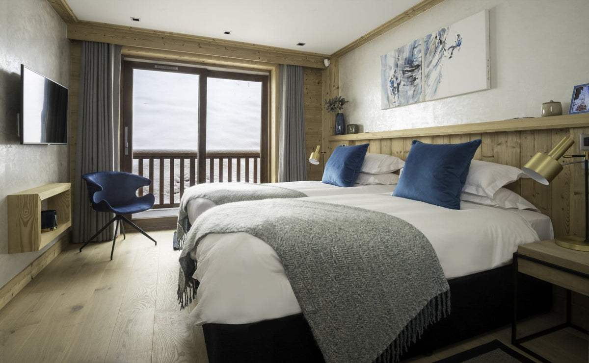 A modern ski resort bedroom with two single beds, white bedding, blue pillows, a gray throw blanket, light wood walls and floor, a blue chair, and a large window overlooking a snowy mountain view.