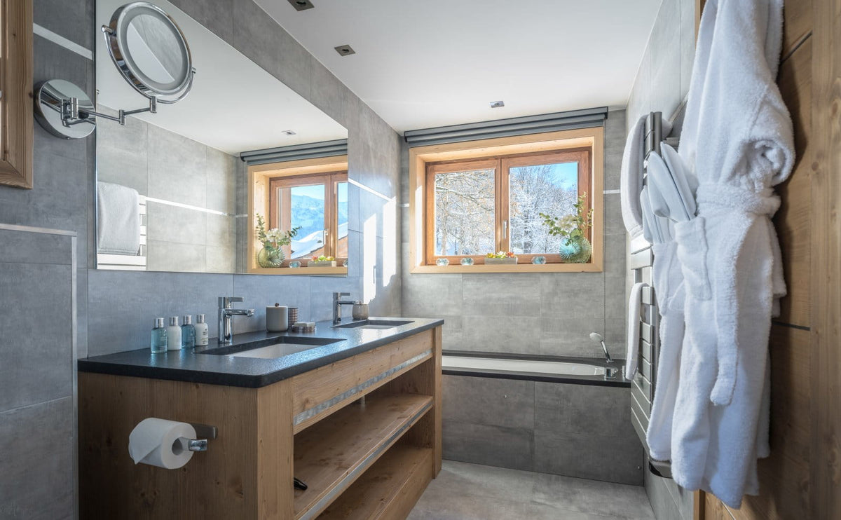 A modern luxury bathroom featuring a double-sink vanity with a dark countertop and open wood shelving, a large wall-mounted mirror with an adjustable magnifying lens, gray stone tile walls and floor, a bathtub by a window, and white bathrobes hanging on a wooden cabinet.