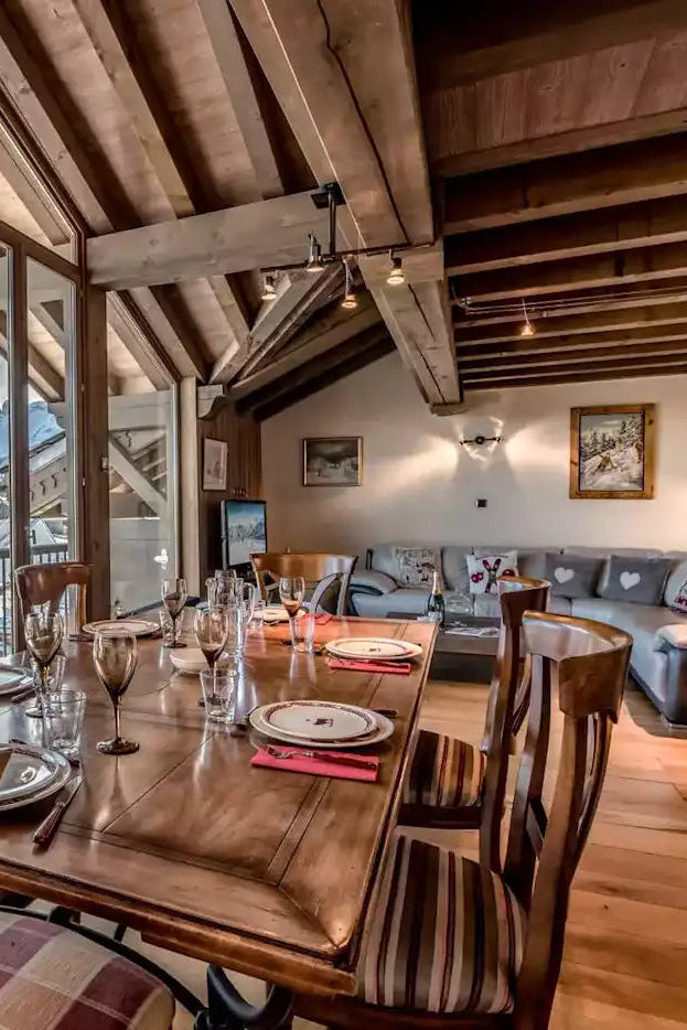 A luxury chalet-style dining room featuring a wooden dining table and chairs set with glassware, overlooking a snowy mountain valley. The background reveals a modern open-plan living room with a gray sectional sofa, wooden ceiling beams, and large floor-to-ceiling windows.