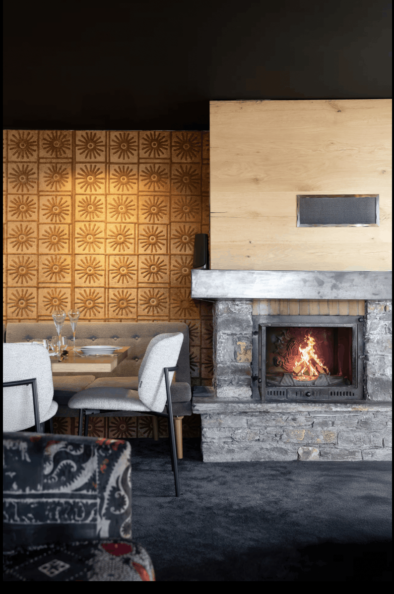 A cozy interior scene featuring a stone fireplace with a glowing fire, a light wood mantel, a patterned orange-brown tiled wall, upholstered dining chairs, and a dark textured floor.