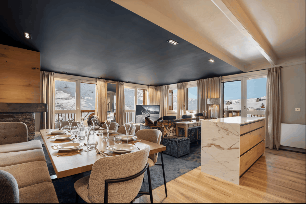 A luxury modern chalet interior featuring a dining area with a wooden table set for six, beige upholstered dining chairs, a marble-topped kitchen island, and large windows overlooking a snowy mountain landscape.