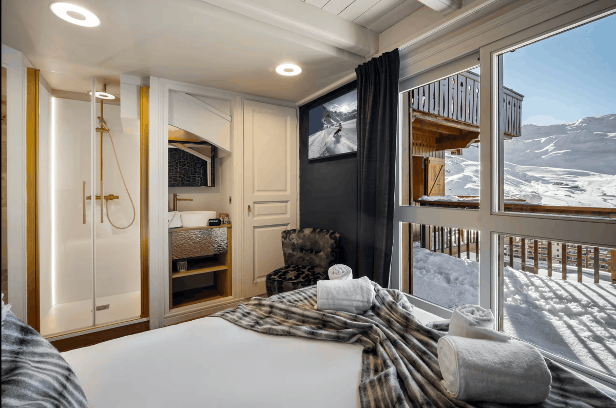 A luxury ski chalet bedroom featuring a large bed with white linens and a plaid throw, a walk-in closet, a glass shower enclosure, and floor-to-ceiling windows overlooking a snowy mountain landscape.