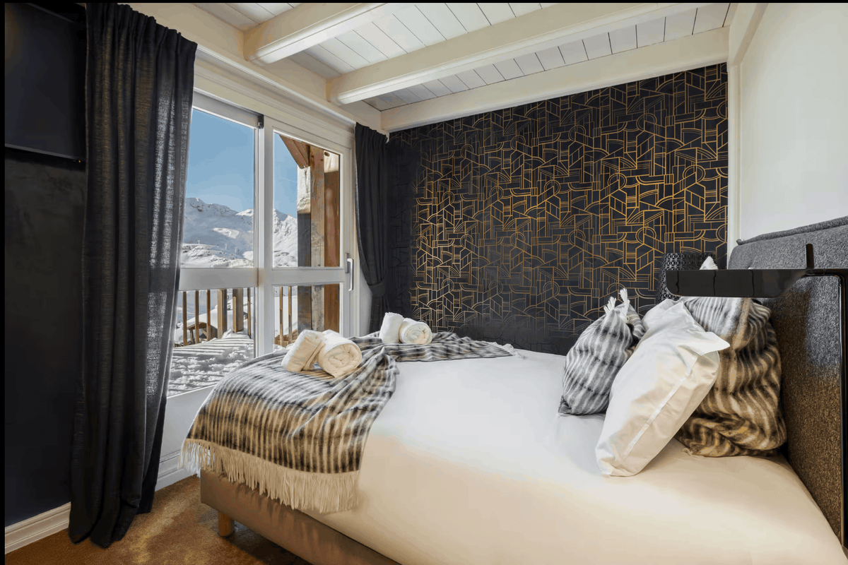A modern bedroom featuring a large bed with white linens, striped gray and beige throw blankets, and patterned pillows, set against a dark wall with gold geometric lines. A large window with dark curtains offers a view of snowy mountains outside.