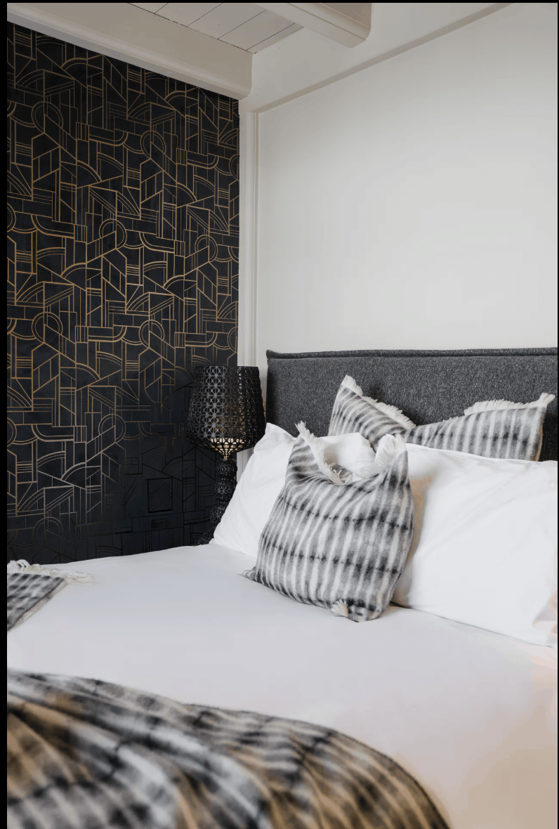 A modern bedroom scene featuring a bed with white linens, striped gray and white pillows, and a textured dark gray headboard. A prominent black geometric patterned accent wall is on the left, with a black patterned table lamp placed on a nightstand.