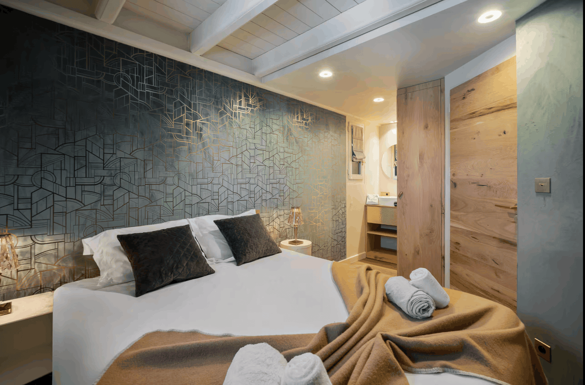 A modern bedroom interior featuring a large bed with white bedding and tan throw blanket, set against a textured blue-gray geometric accent wall and a light wood door.