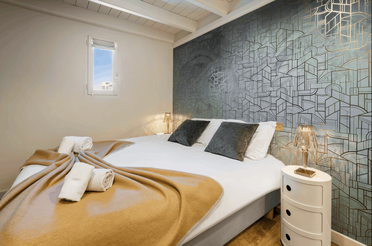 A modern bedroom featuring a bed dressed in white linens and a tan blanket, set against a distinctive geometric patterned accent wall in blue-gray tones. The room includes a white nightstand, a window, and warm ambient lighting.