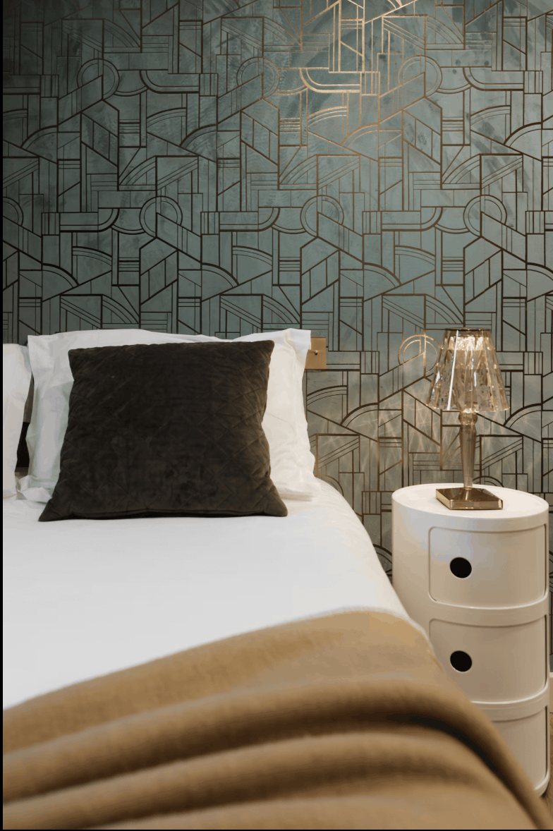 A styled bedroom corner featuring a bed with white bedding and a dark brown accent pillow, next to a white nightstand with a glass table lamp. The wall behind is covered in a textured geometric wallpaper with a muted teal-green background and gold line art.