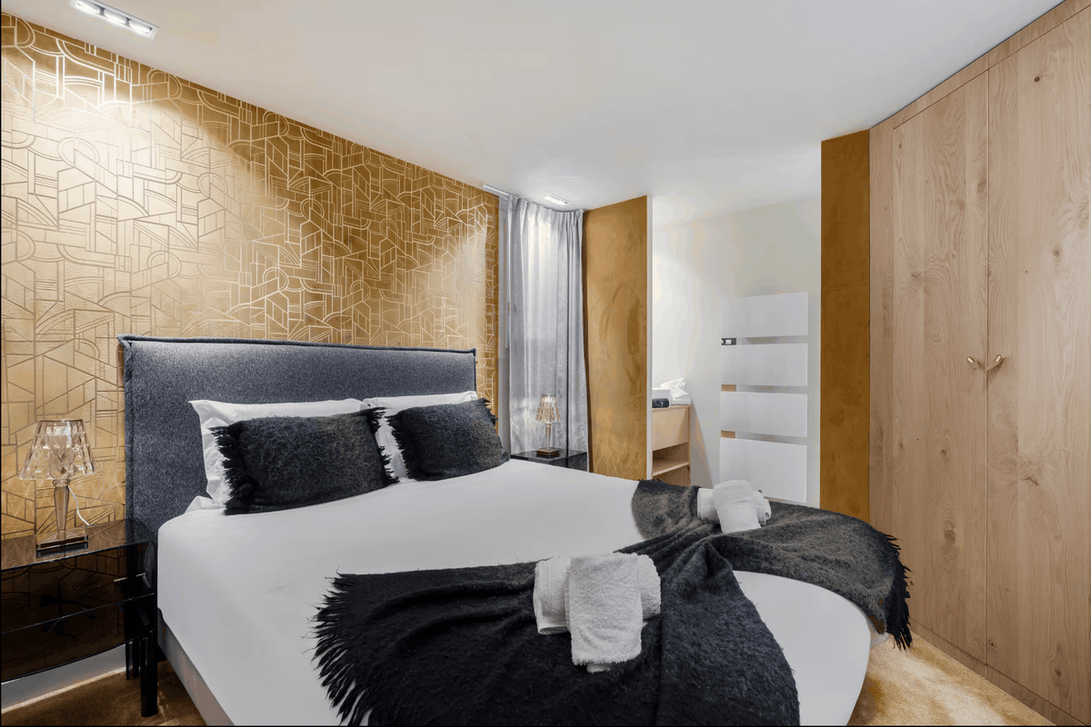 A modern bedroom featuring a bed with white linens and dark gray pillows and throw, set against a geometric gold wallpaper accent wall and light wood paneling.