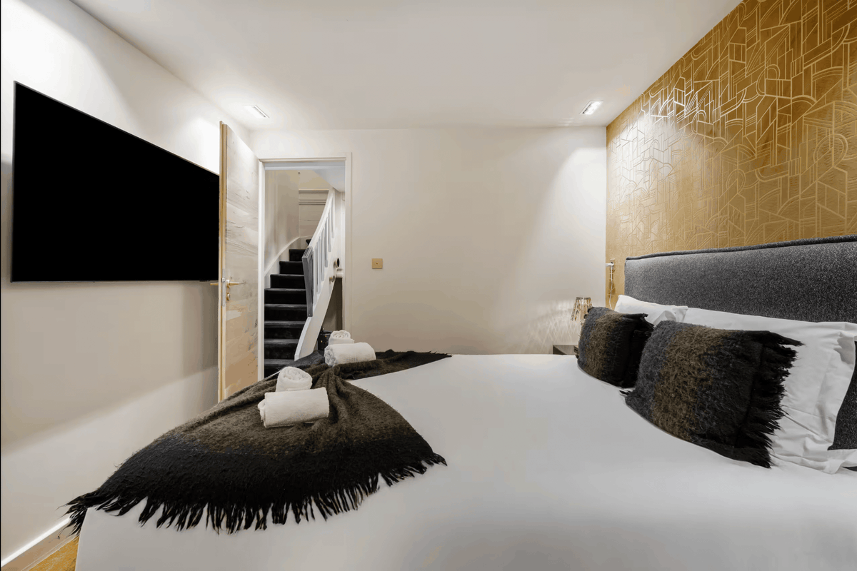 A modern bedroom interior featuring a large bed with white bedding, black textured pillows, and a dark fringed throw blanket. The wall behind the headboard has a gold geometric pattern, and a staircase is visible in the background through an open doorway.
