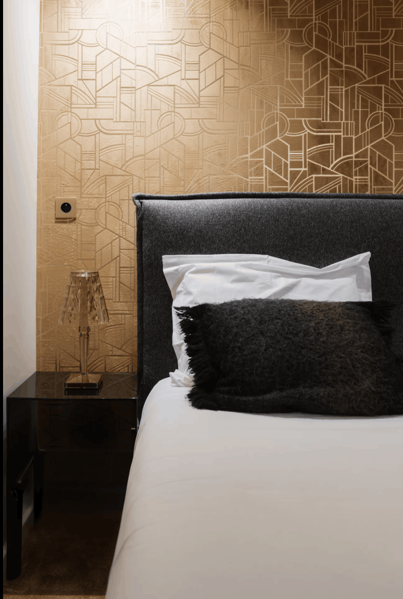 A modern bedroom corner featuring a bed with white bedding, a dark textured headboard, a black bedside table with a decorative lamp, and a prominent geometric patterned wall in warm beige and gold tones.