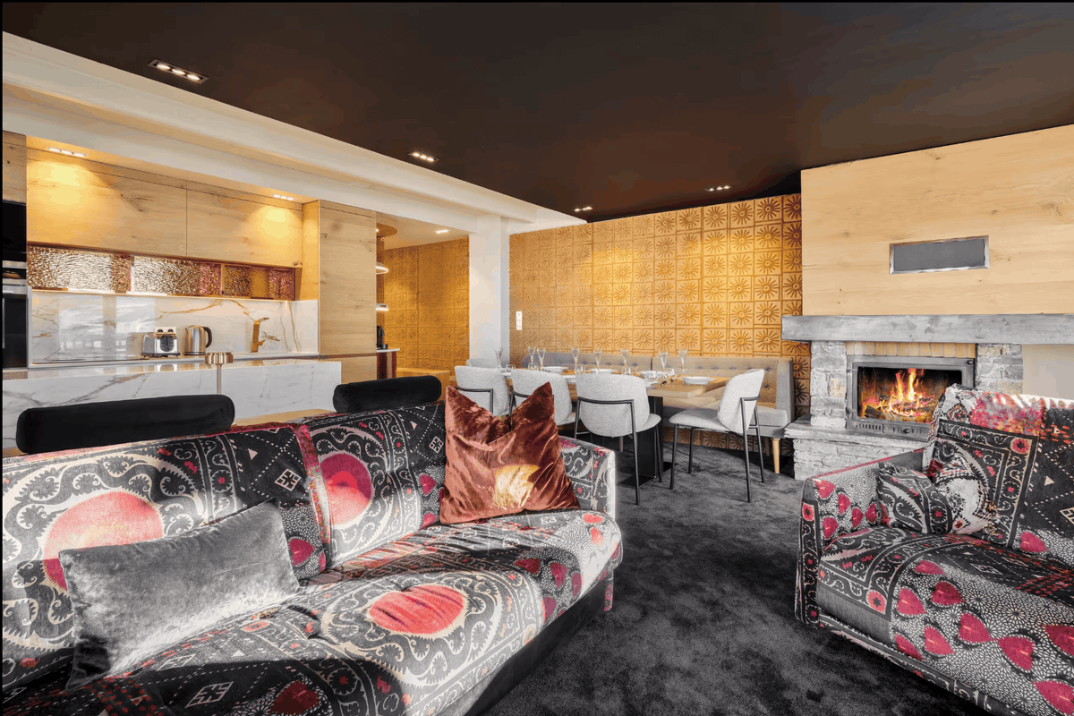 A luxury modern living room featuring a patterned black velvet sofa with red and gray accents, a matching armchair, a dark gray shag carpet, a wood-paneled fireplace, and a dining area in the background.