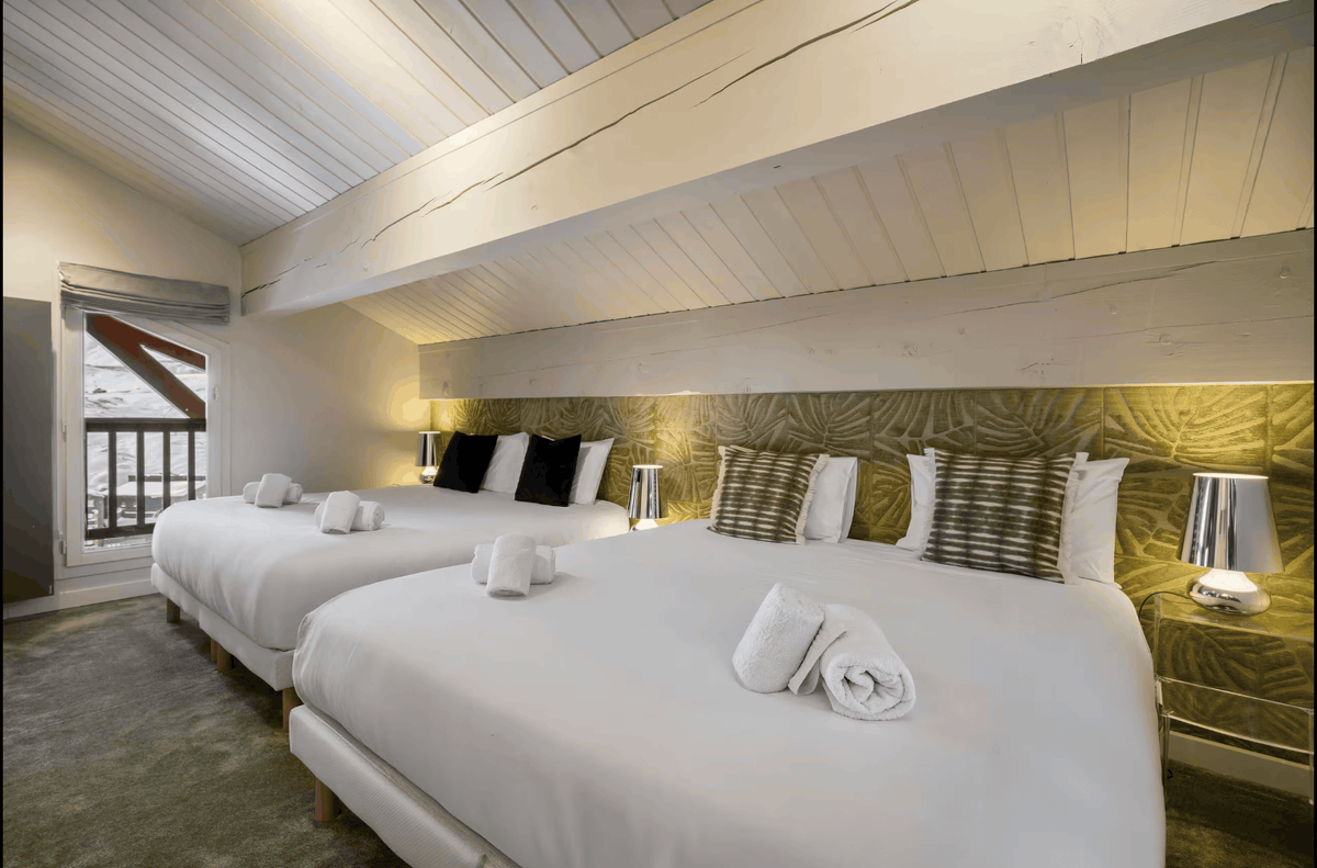 A luxury mountain-style bedroom featuring two neatly made beds with white linens, decorative pillows, and folded towels, set under a sloped white wood ceiling with a warm yellow backlit wall panel.