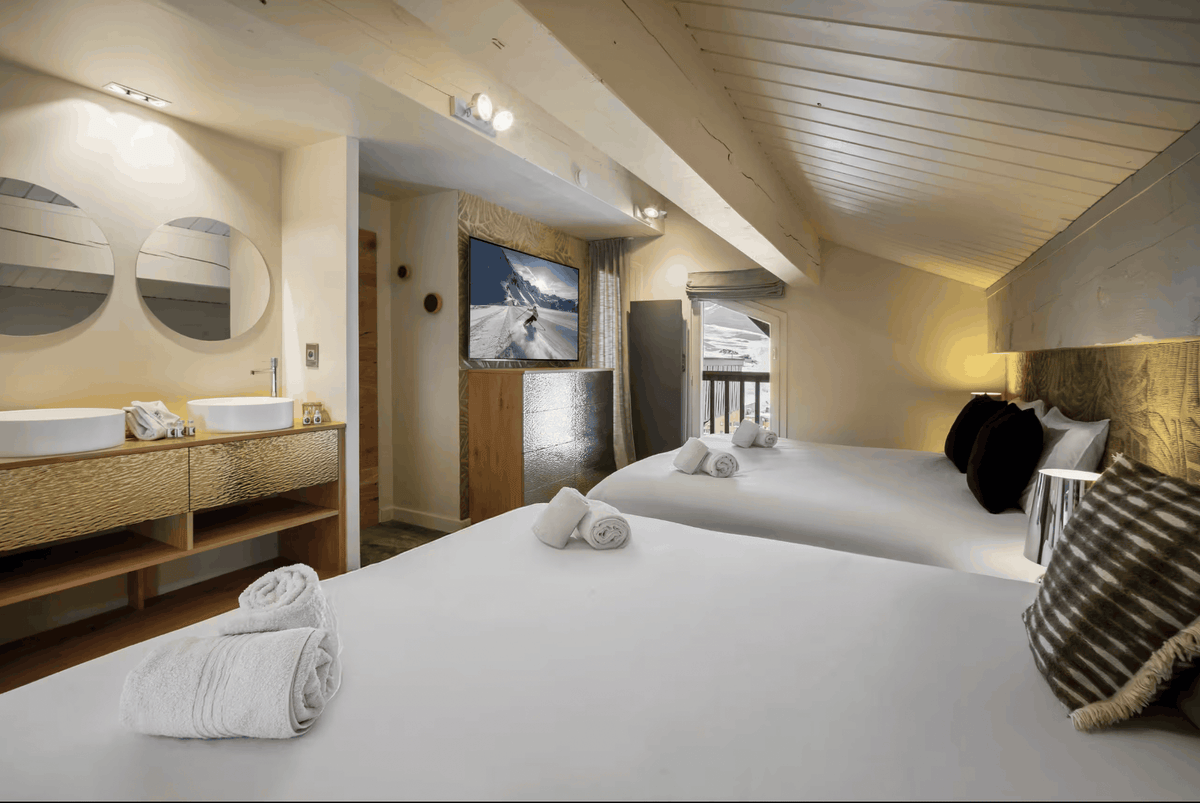 A luxury ski resort hotel room featuring two double beds with white linens and dark pillows, a modern wood vanity with double sinks, and a warm wood-paneled ceiling.