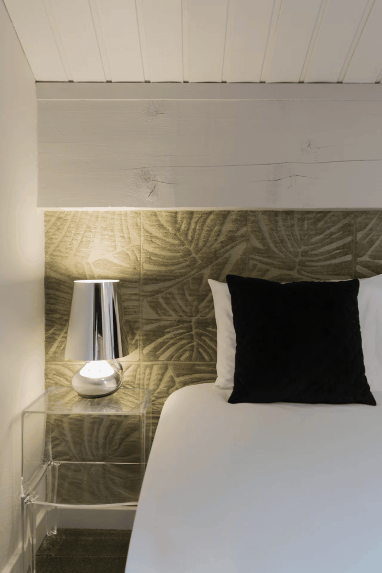 A modern bedroom corner featuring a bed with white bedding and a black accent pillow, set against a textured olive green leaf-patterned headboard and a clear glass side table with a silver lamp.