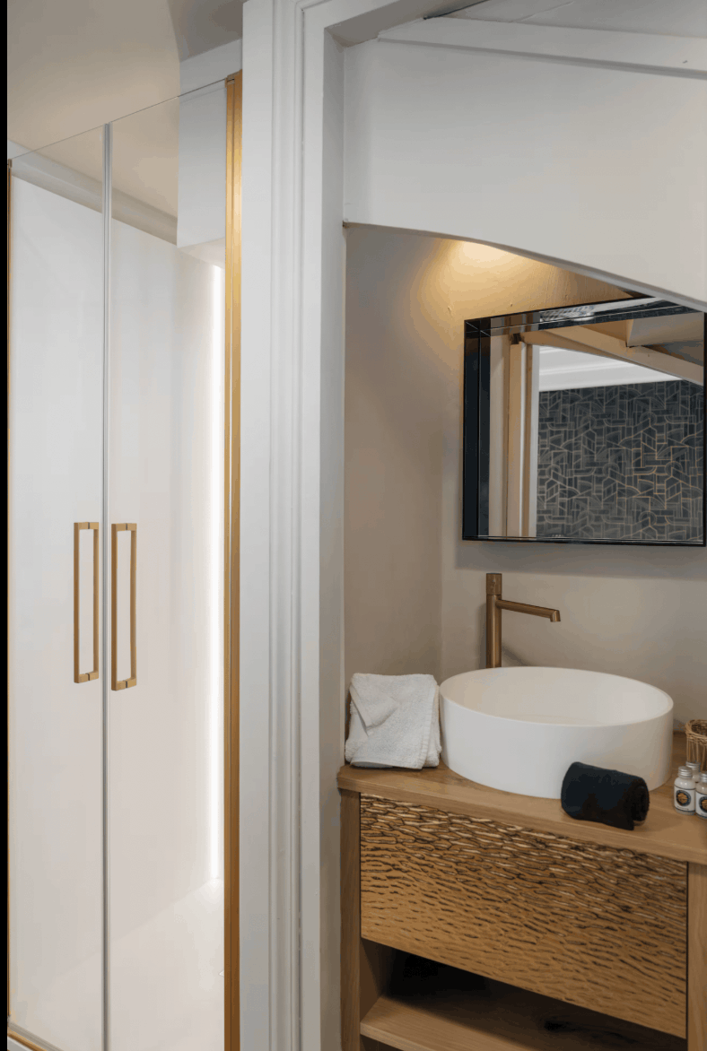A modern bathroom interior featuring a white vessel sink on a textured wood vanity, a gold faucet, a wall mirror, and a white wardrobe with gold handles in the adjacent room.