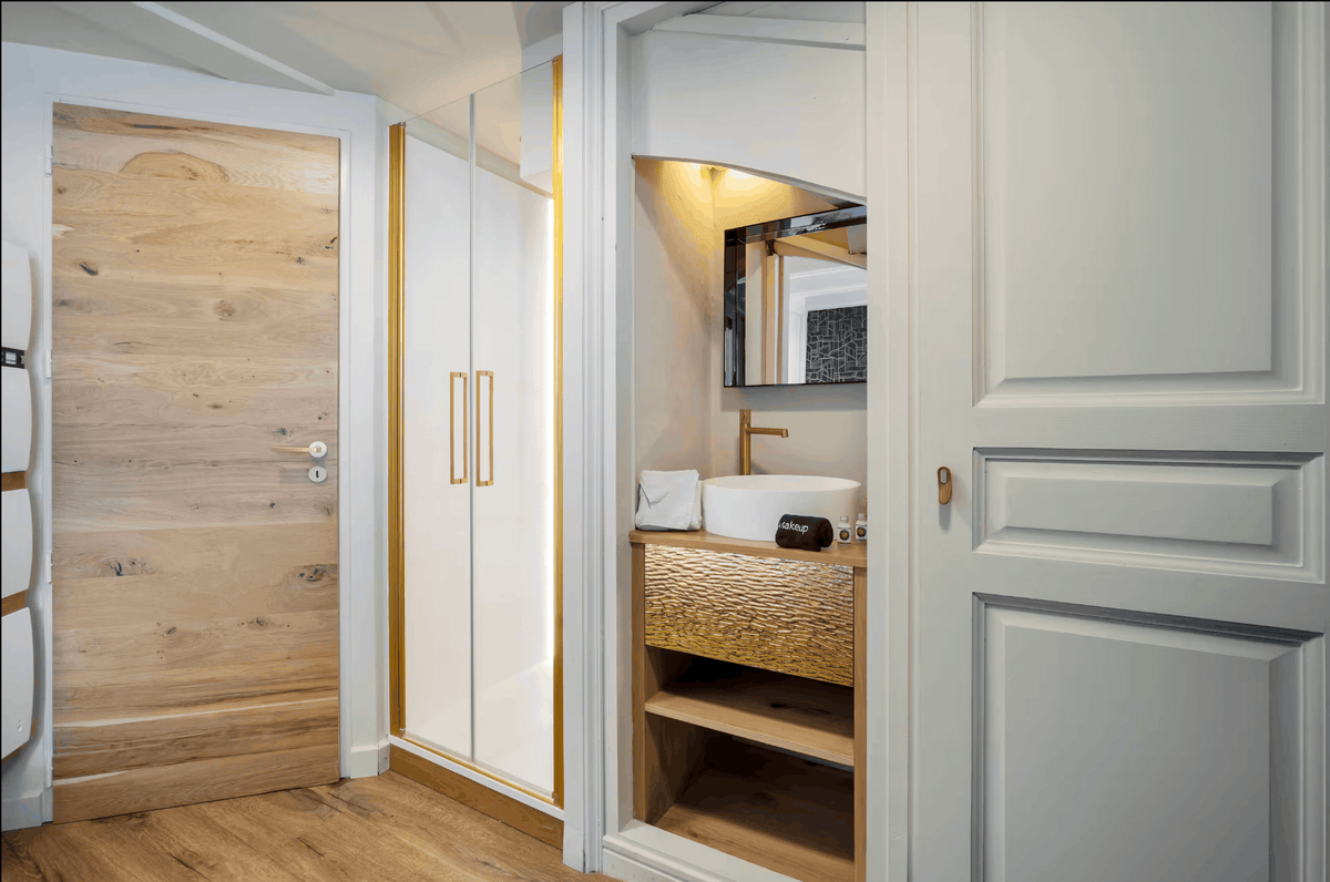 A modern interior entryway featuring a light wood paneled door on the left, a white wardrobe with gold handles in the center, and an open doorway on the right showing a bathroom vanity with a white sink, textured gold cabinet front, and mirror.