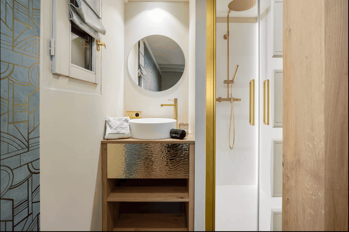 A modern bathroom vanity with a white vessel sink, textured gold cabinet front, round wall mirror, and matching gold faucet. A glass shower door with gold trim and a white shower area are visible to the right.