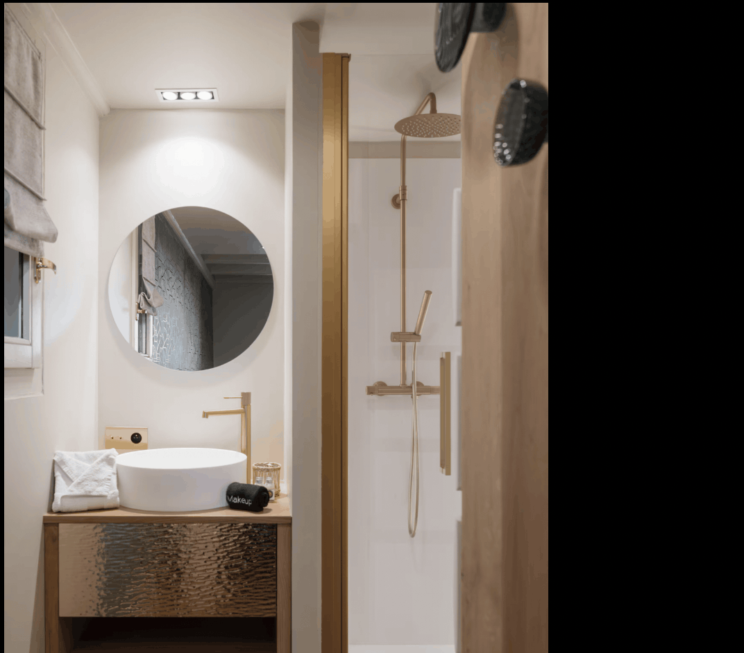 A modern bathroom interior featuring a vessel sink with a hammered gold vanity cabinet, a round wall mirror, and a walk-in shower with matching gold fixtures.