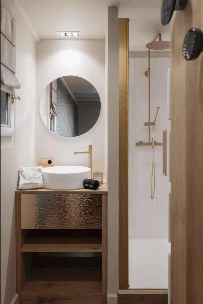 A modern bathroom interior featuring a wooden vanity with a white vessel sink, a round wall mirror, and a textured metallic drawer front. A walk-in shower area with warm brass fixtures is visible to the right.