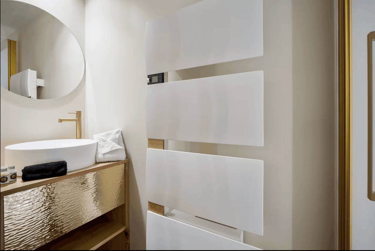 A modern bathroom interior featuring a white vessel sink on a textured gold vanity, a round wall mirror, and a tall white cabinet with horizontal gold-framed panels.