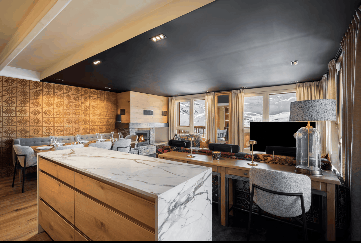 A luxury modern ski chalet interior featuring a large open-plan kitchen and dining area with a marble-topped island, wood cabinetry, a dark ceiling, patterned gold wall panels, and large windows overlooking a snowy mountain landscape.