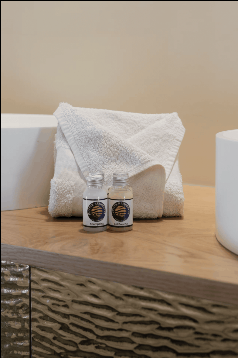 Two small toiletry bottles with white labels and silver caps sit on a wooden vanity counter in front of a folded white towel. The setting appears to be a bathroom or vanity area with a beige wall and part of a white sink visible.