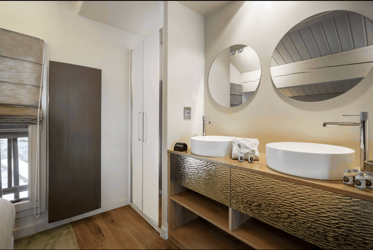 A modern bathroom vanity featuring two white oval vessel sinks on a textured metallic gold-foiled wooden cabinet with a light wood countertop. Above the sinks are two round mirrors, and the space is illuminated by warm overhead lighting.