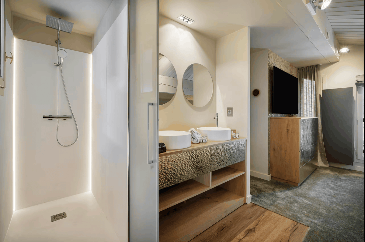 A modern bathroom interior with a walk-in shower on the left, a double vessel sink vanity with round mirrors in the center, and a television mounted above a wooden cabinet on the right.