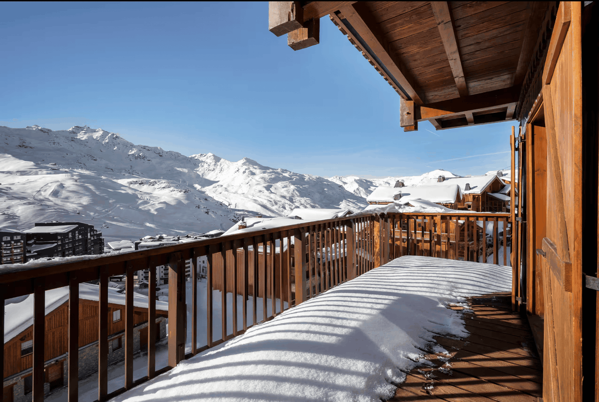 A sunny wooden balcony overlooking a snowy mountain village under a clear blue sky, with snow-covered peaks and chalets in the background.