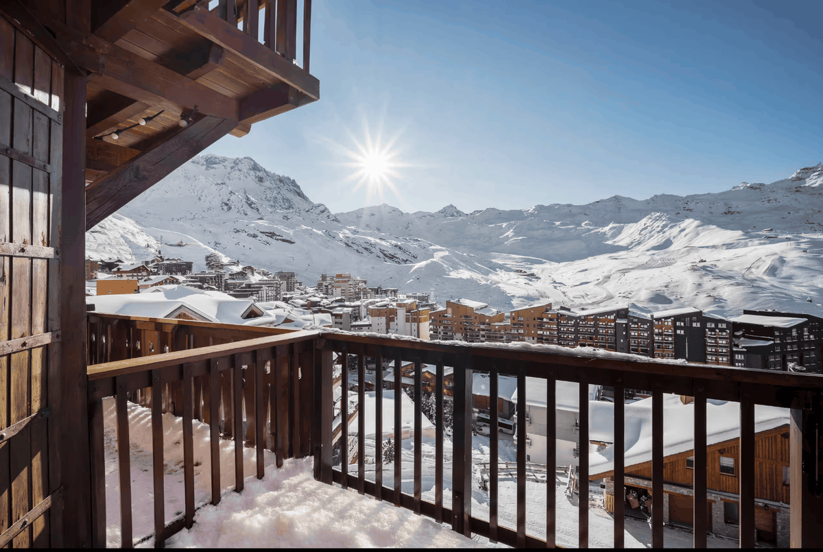 A scenic winter mountain view from a wooden balcony, with snow-covered peaks, a small village, and a bright sun in a clear blue sky.