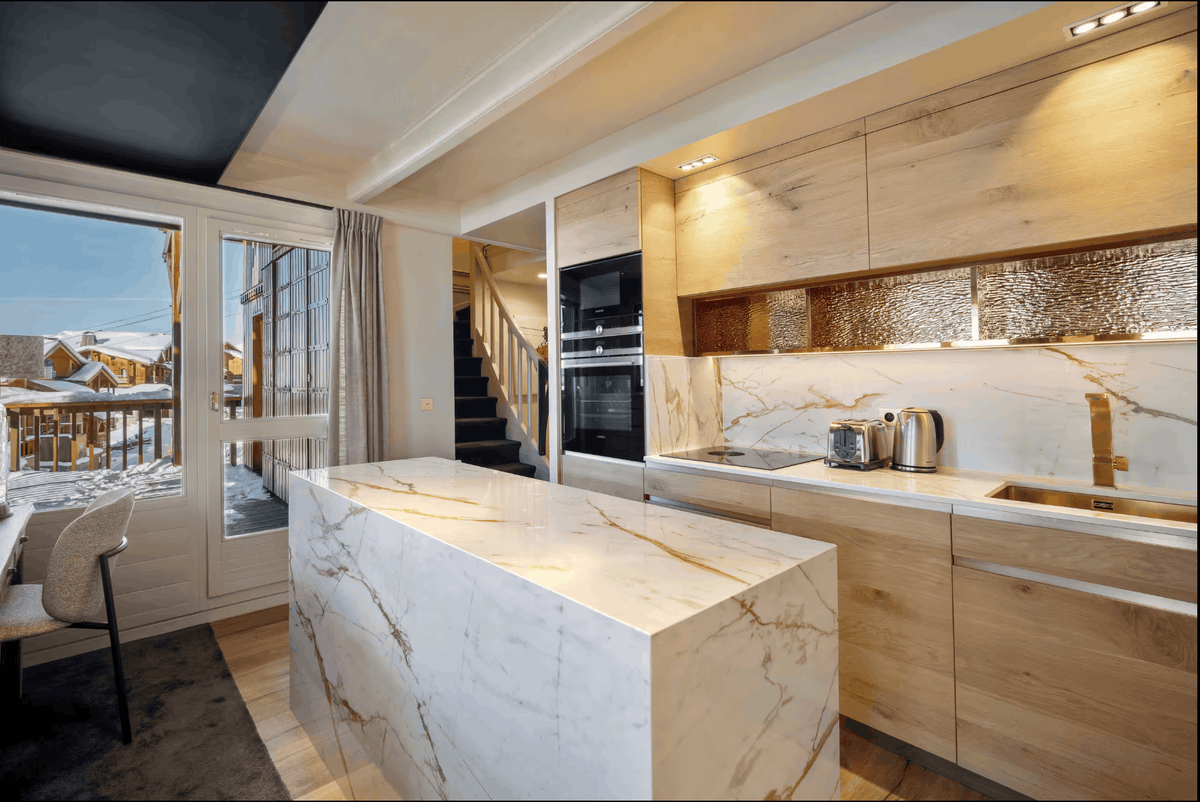 Modern kitchen interior with a large white marble island, light wood cabinetry, built-in black appliances, and a textured metallic backsplash. Snow-covered roofs and outdoor windows are visible in the background.
