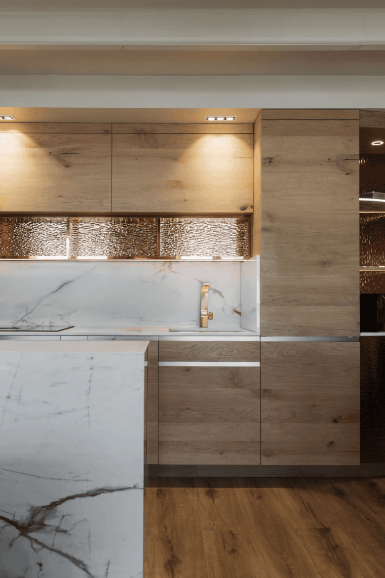Modern kitchen cabinetry featuring light oak wood cabinets, a white marble countertop and backsplash, and a hammered metallic copper backsplash panel.