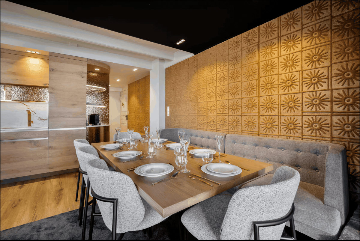 A modern dining room setup featuring a rectangular wooden table set with white plates and glassware, surrounded by gray upholstered chairs. The dining area is set against a prominent wall with gold sunburst-patterned wall panels and a tufted gray bench.