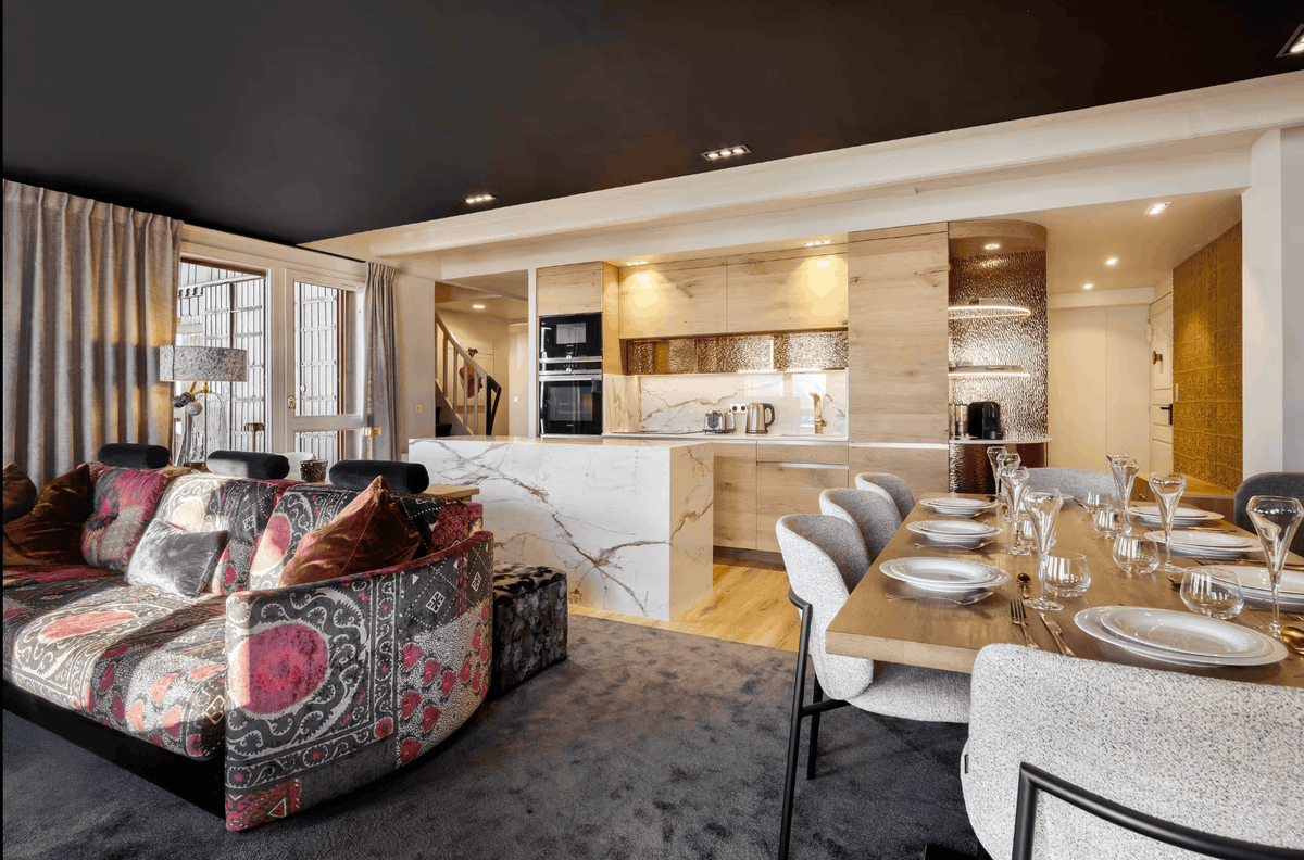 A luxury modern open-plan living and dining space featuring a patterned velvet sofa in the foreground, a marble kitchen island, and a dining table set with glassware under warm lighting.