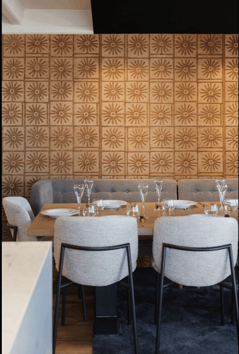 A dining area with a rectangular wooden table set with white plates and clear glassware, surrounded by light gray upholstered chairs with black metal frames. A tufted gray banquette sits against a wall covered in warm brown sunburst-patterned tiles.
